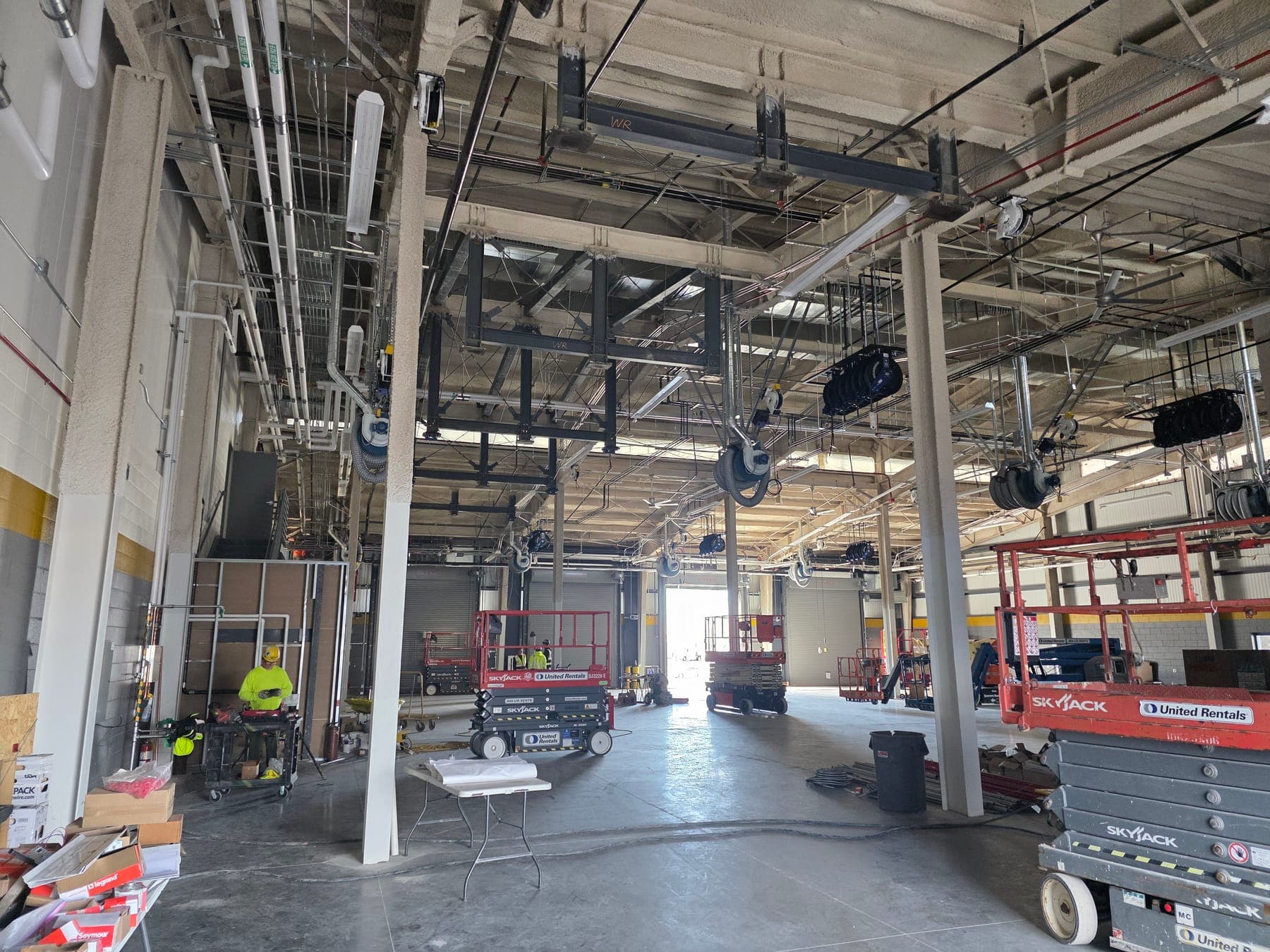 Maintenance bay interior during construction, with overhead hose reels, piping and conduit, roll-up doors, and scissor lifts on the floor
