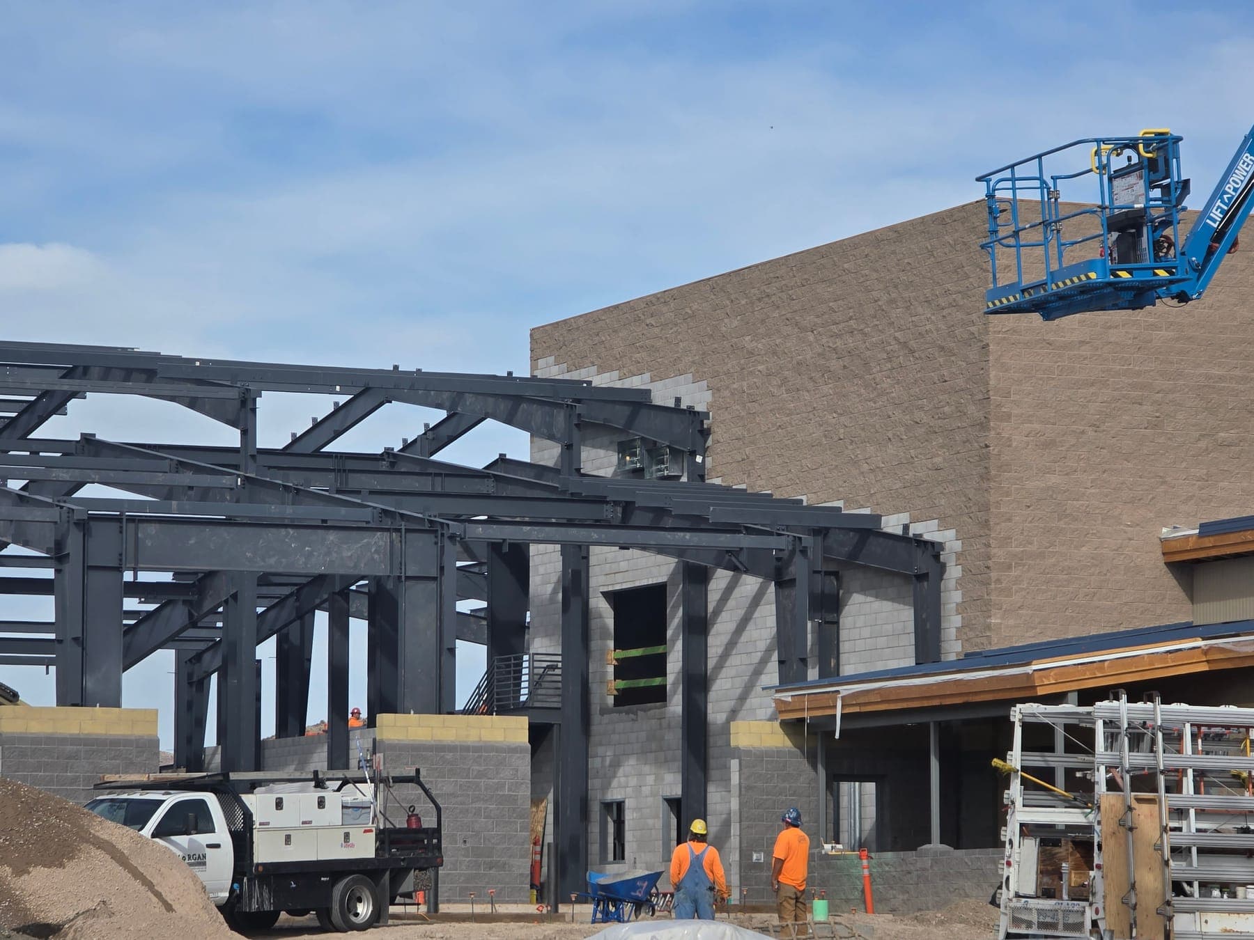 Two workers in hard hats looking up at erected steel building frames next to a masonry wall, with a work truck and a boom lift on site