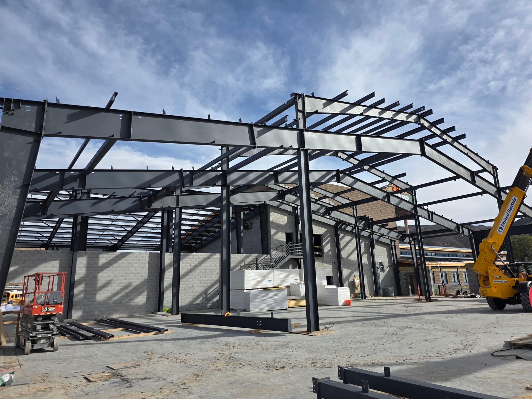 Dark steel building frames with roof purlins erected on a concrete slab beside a masonry building, with a telehandler on the right