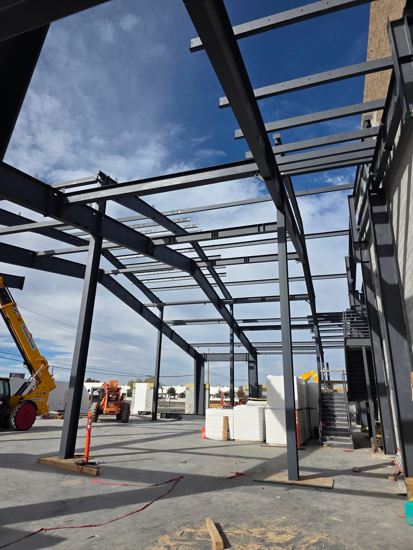 Looking up through erected steel columns, beams, and roof purlins toward a blue sky, with a steel stair and stacked insulation panels on the slab