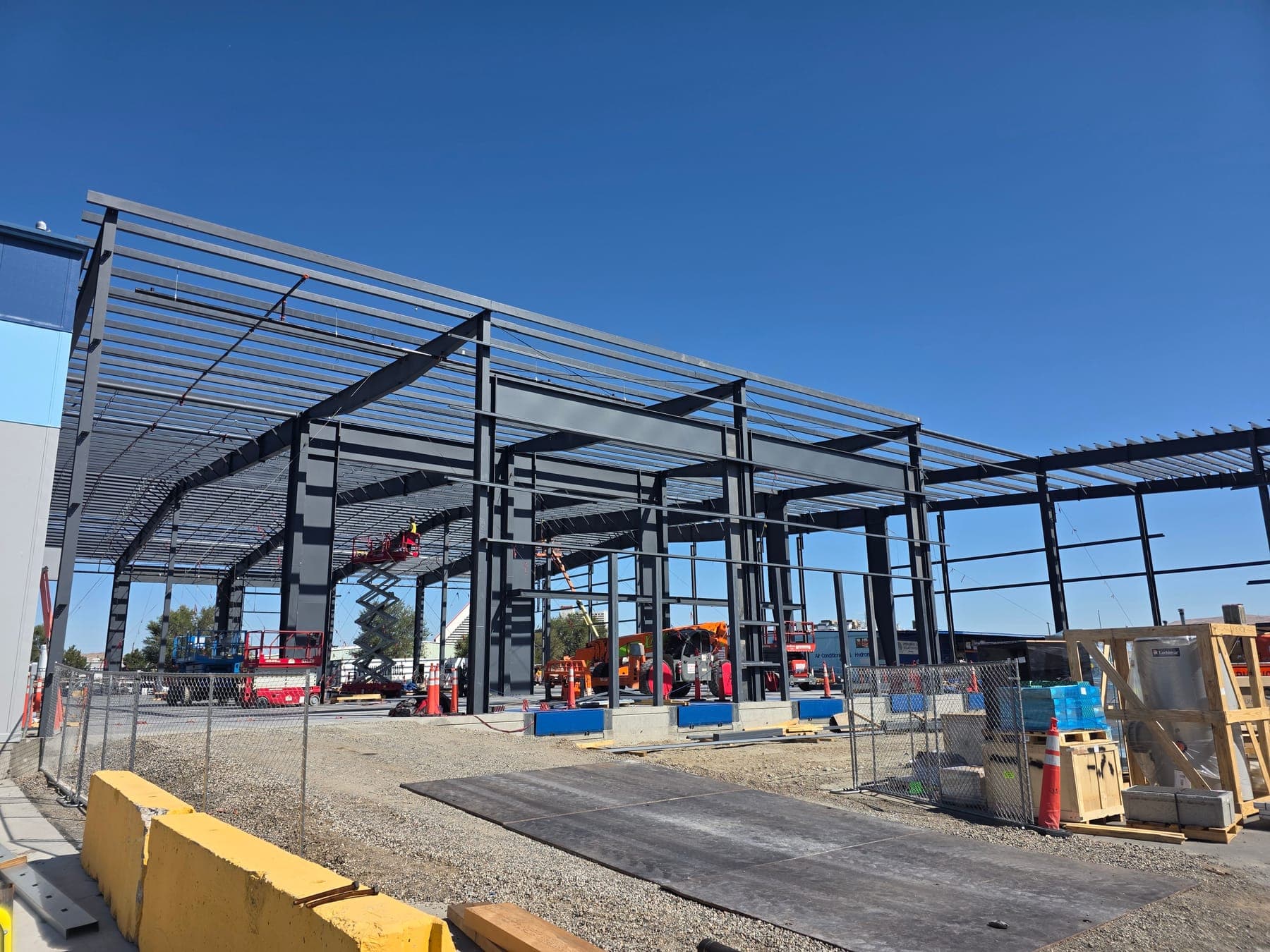 Steel rigid frames, roof purlins, and wall girts of a metal building under erection next to an existing building, with lifts on site